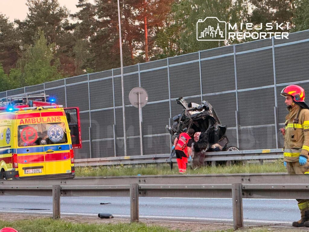Ratownicy medyczni udzielają pomocy osobie w przewróconym samochodzie na drodze, obok stoi strażak.