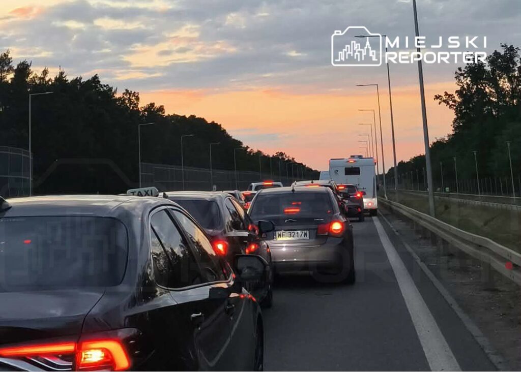 Ruch samochodowy w korku na drodze, w tle zachód słońca i zieleń drzew.
