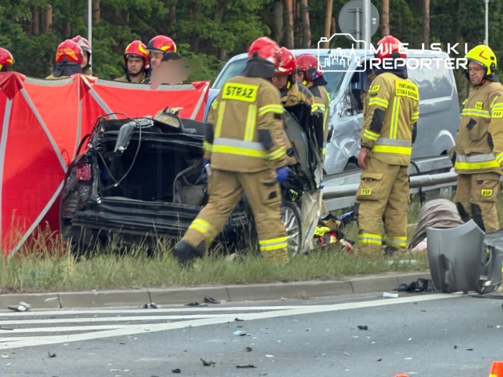 Strażacy w mundurach pracują przy wraku samochodu osobowego na drodze, otoczeni przez czerwony namiot.