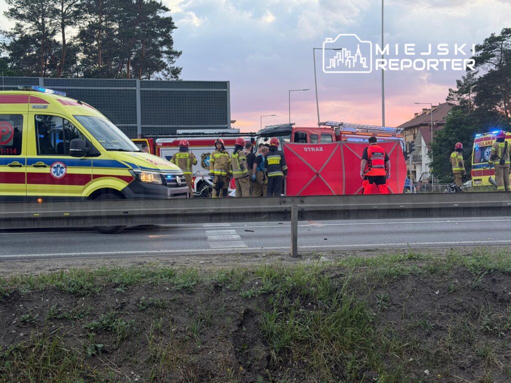 Grupa ratowników i strażaków pracuje przy przewróconym pojeździe na drodze, obok zaparkowanej karetki.