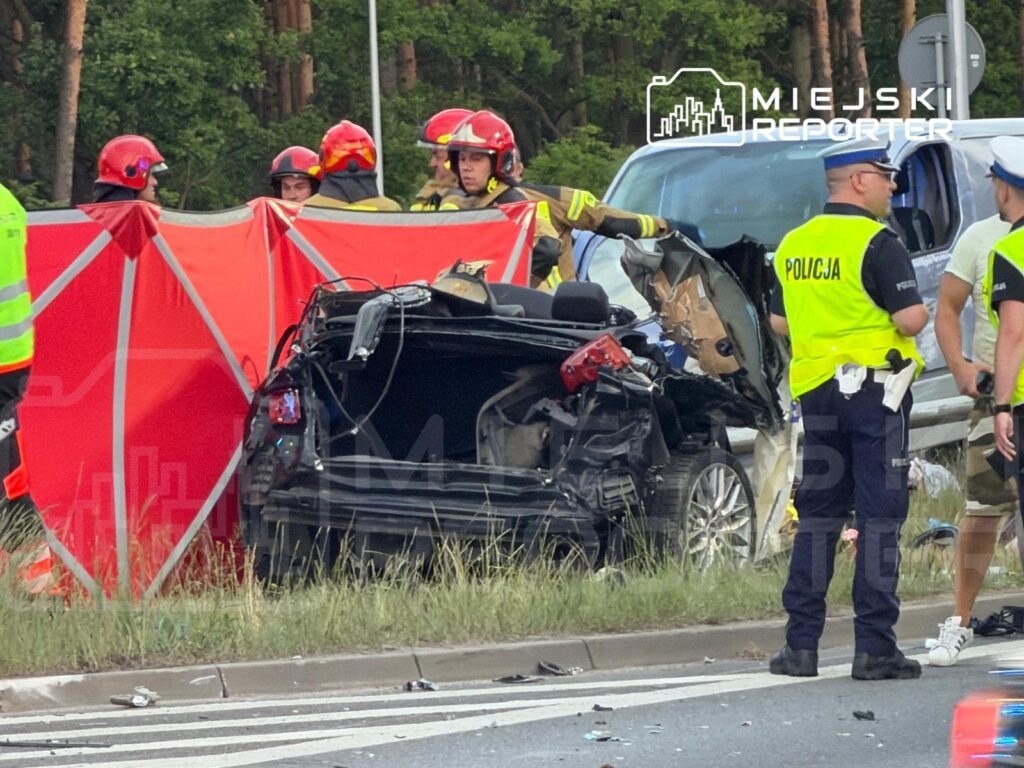 Grupa strażaków i policjantów pracuje przy rozbitej, czarnej osobówce na drodze, otoczonej czerwonym parawanem.