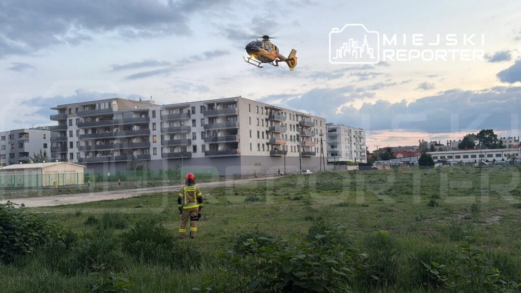Śmigłowiec ratunkowy przelatuje nad terenem zielonym, obok stoi strażak w mundurze oraz nowoczesne budynki mieszkalne w tle.