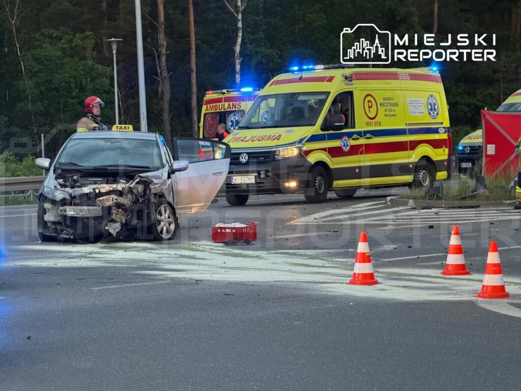 Uszkodzony samochód osobowy z rozbitym przodem stoi na drodze, obok ratownicy medyczni i karetka.