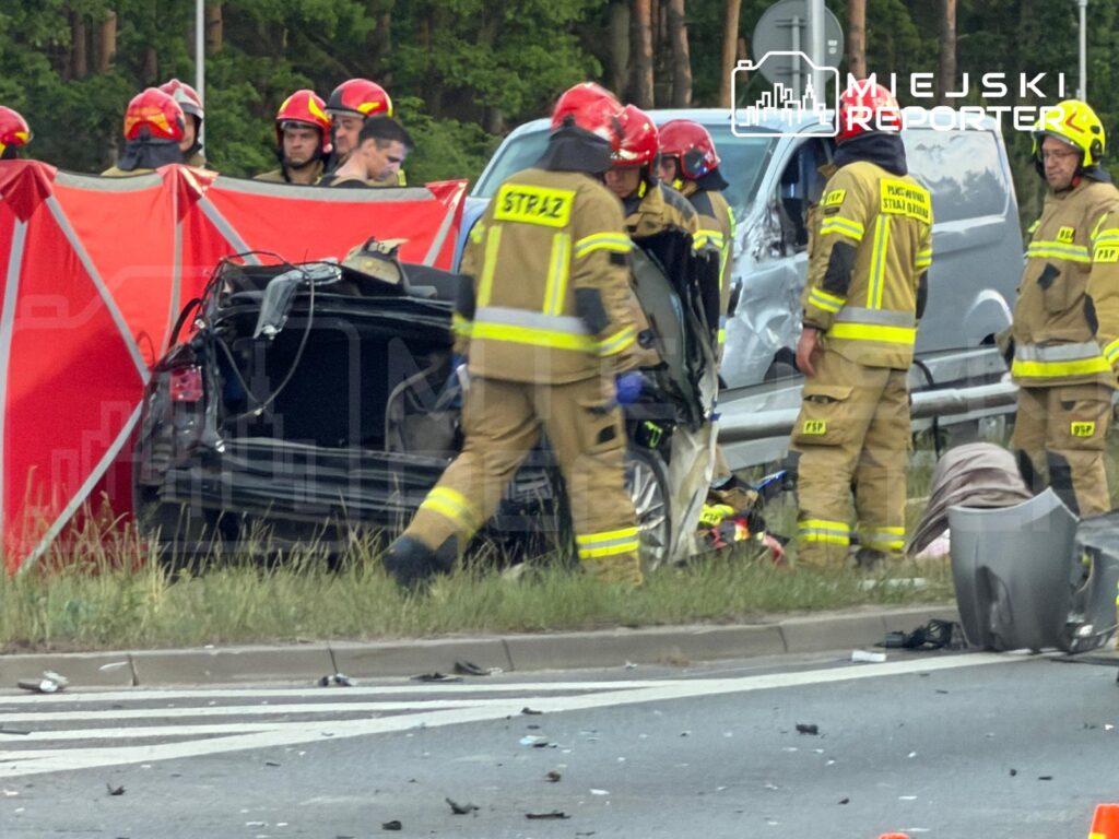 Strażacy w mundurach ratują poszkodowanych z rozbitego samochodu przy drodze, otoczeni przez czerwone namioty.