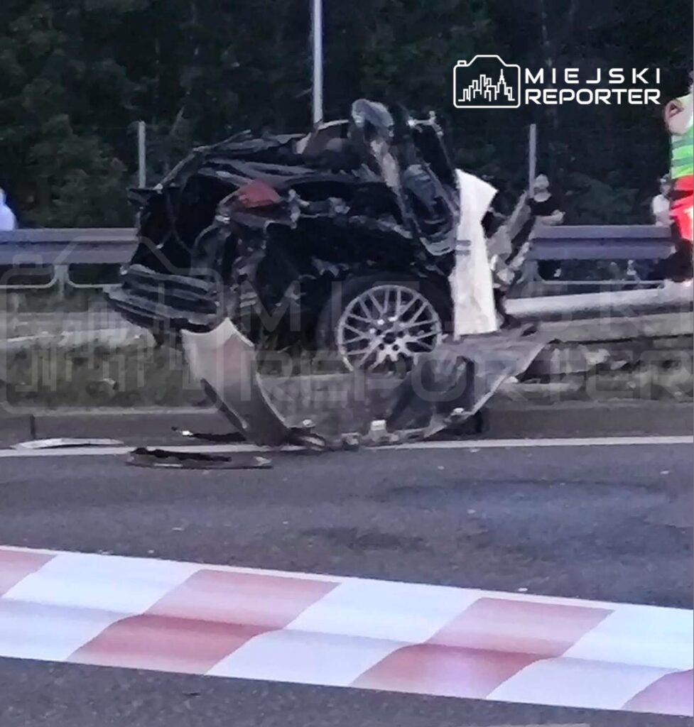 Zniszczony samochód osobowy leży na boku na jezdni, obok widać pasy drogowe i osoby w odblaskowych kamizelkach.