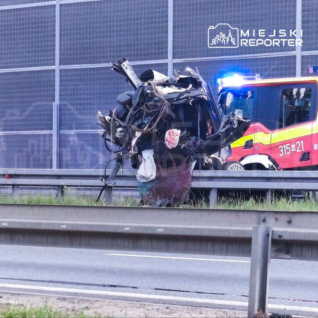 Zniszczony pojazd stoi na poboczu drogi, obok niego widać włączone światła sygnalizacji straży pożarnej.