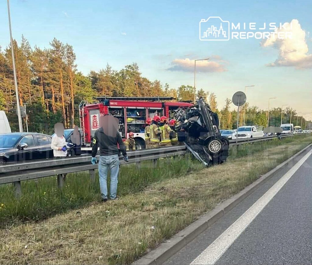 Samochód leżący na boku przy barierze drogowej, obok grupy strażaków i ratowników medycznych w akcji.