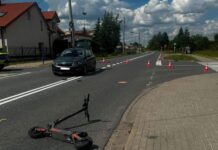Na jezdni leży uszkodzony skuter elektryczny, obok stoi czarny samochód oraz ustawione pachołki drogowe.