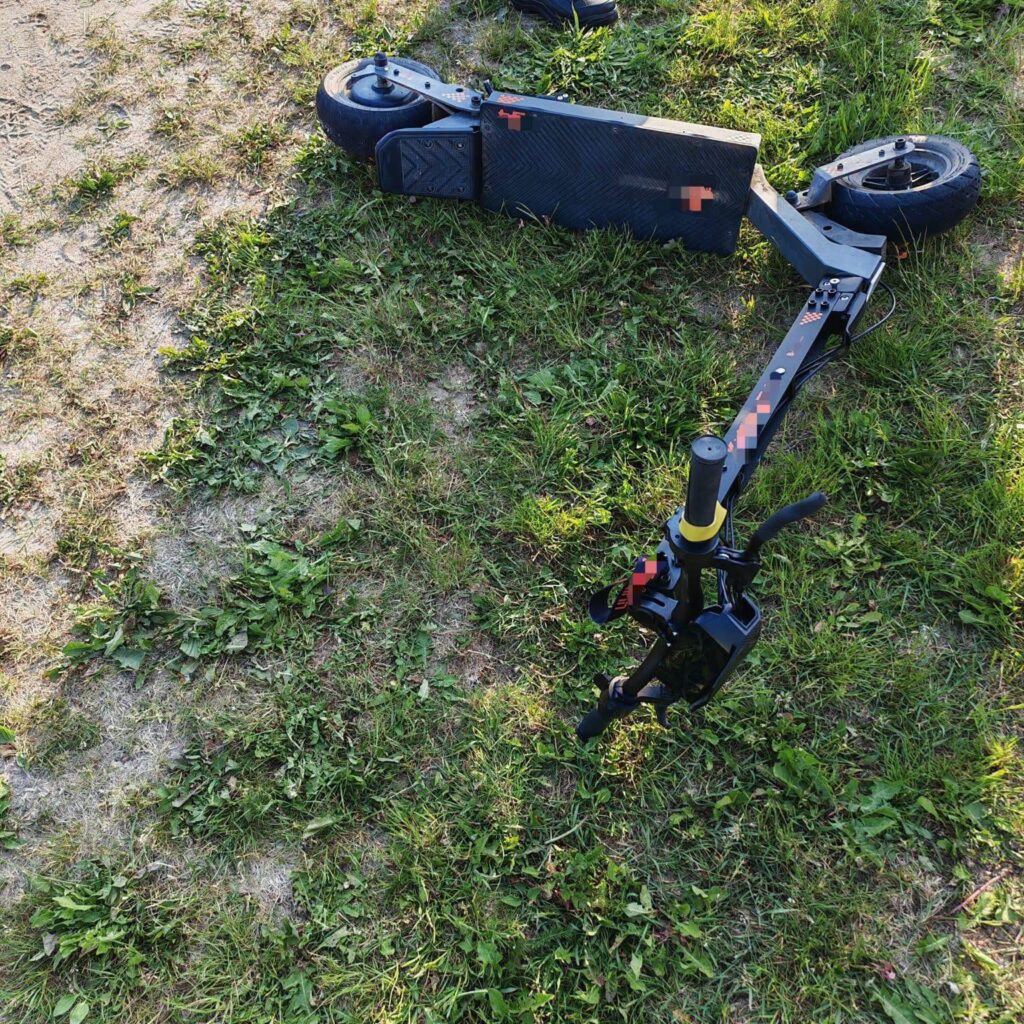 Złamana hulajnoga leży na trawie, obok widoczna jest drobna roślinność i suche miejsca w glebie.