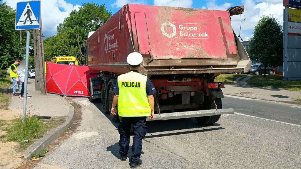 Policjant w kamizelce odblaskowej kieruje ruchem obok przewróconej ciężarówki na drodze. W tle widać karetkę i osoby rozmawiające.