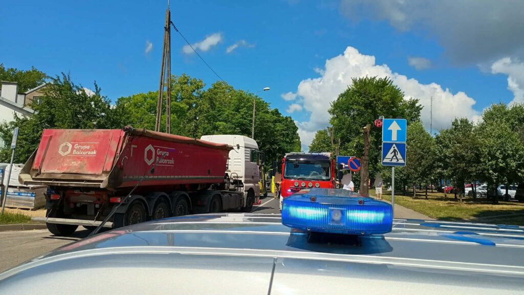 Na drodze stoją pojazdy, w tym czerwona ciężarówka oraz samochód strażacki, otoczone przez zieleń drzew.
