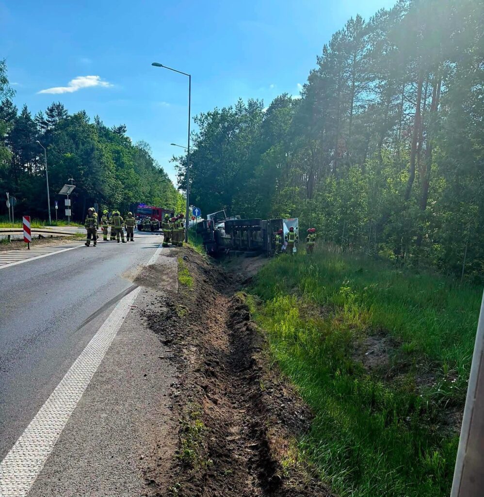 Grupa strażaków pracuje przy przewróconym pojeździe na poboczu drogi, w otoczeniu drzew i zieleni.