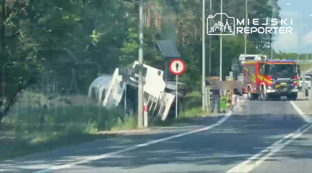 Ciężarówka przewrócona na poboczu drogi, obok straż pożarna i kilka osób obserwujących zdarzenie.