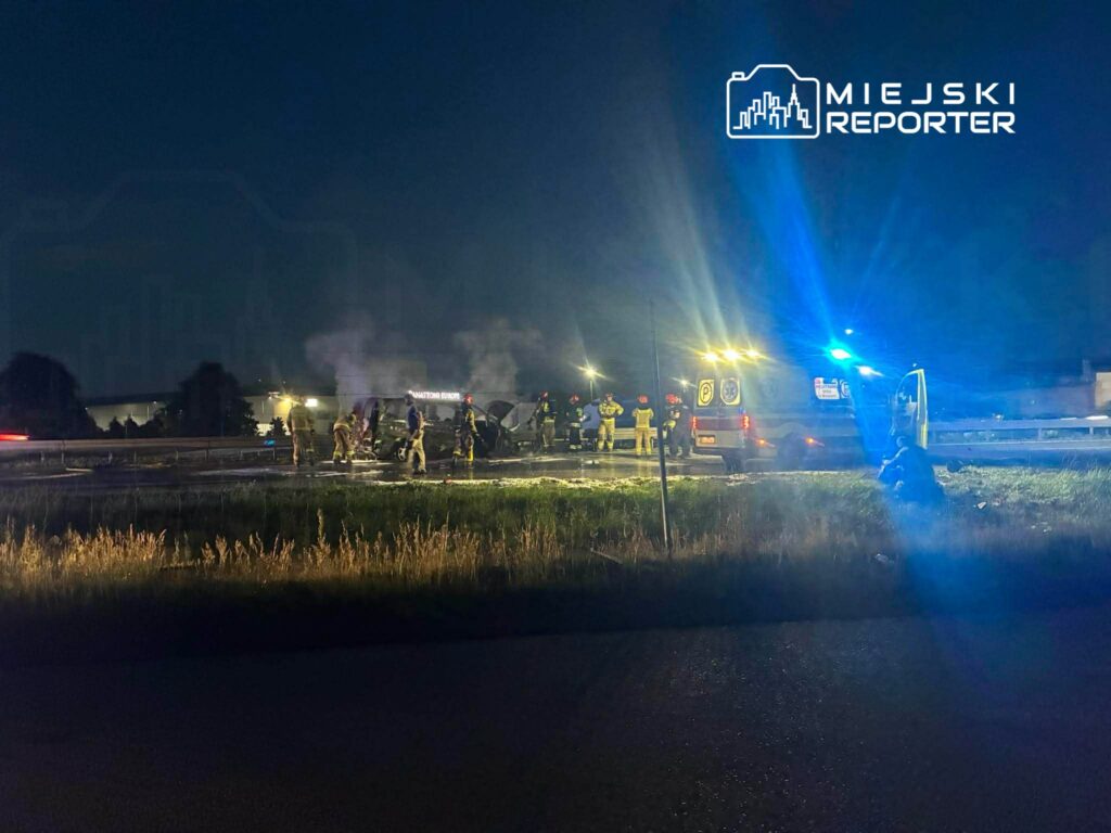 Grupa strażaków gasi pożar pojazdu na drodze, wokół widoczne są kłęby dymu i policyjne światła.