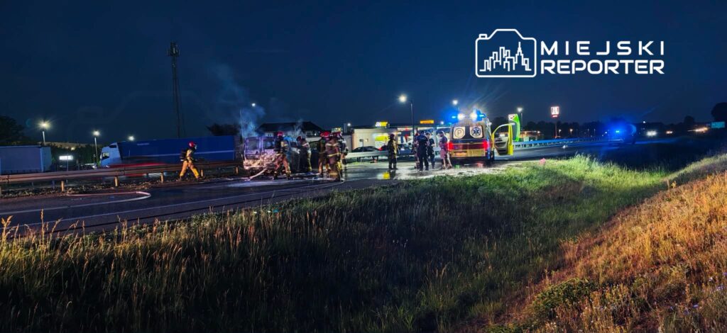 Grupa strażaków gasi płonący pojazd na autostradzie w nocy, obok widać ambulans oraz zaparkowane samochody.