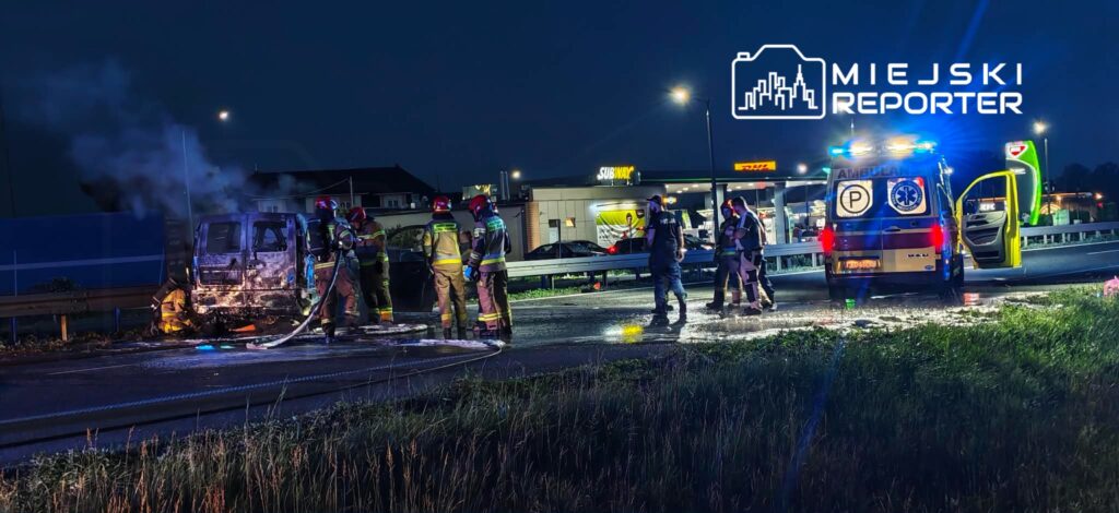 Grupa strażaków gasi płonącą furgonetkę na drodze, obok pojazdów ratunkowych w nocy. W tle widać restauracje i sklepy.