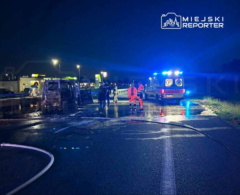 Strażacy gaszą spalone auto na drodze, obok znajdują się ratownicy medyczni i karetka z włączonymi sygnałami.