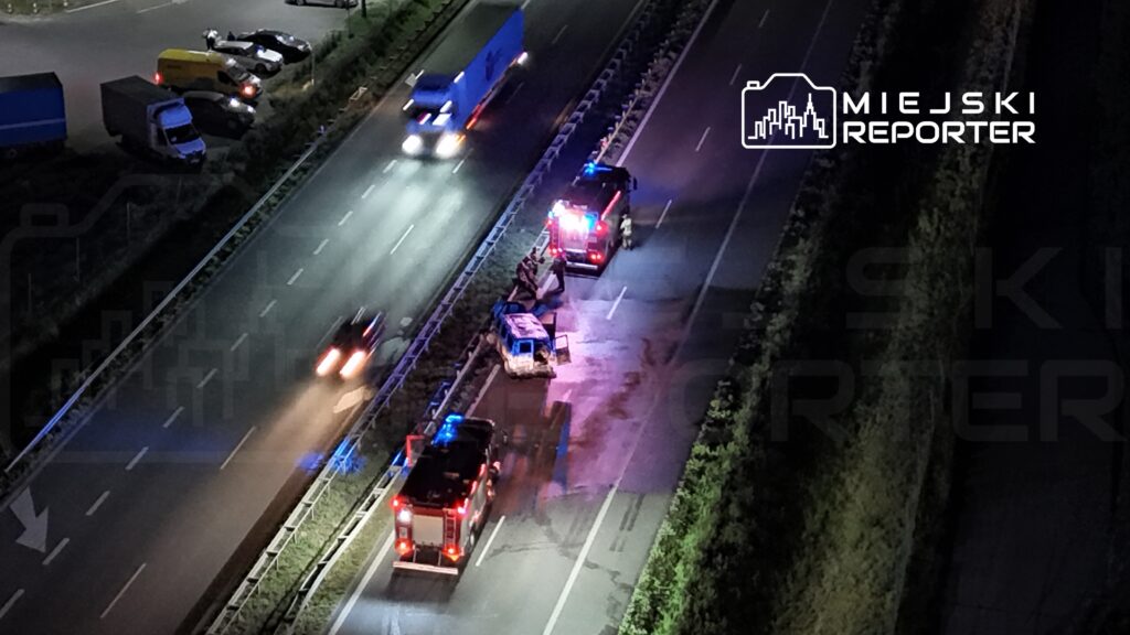 Na drodze krajowej, dwa wozu strażackie z włączonymi sygnałami świetlnymi interweniują przy przewróconym pojeździe.