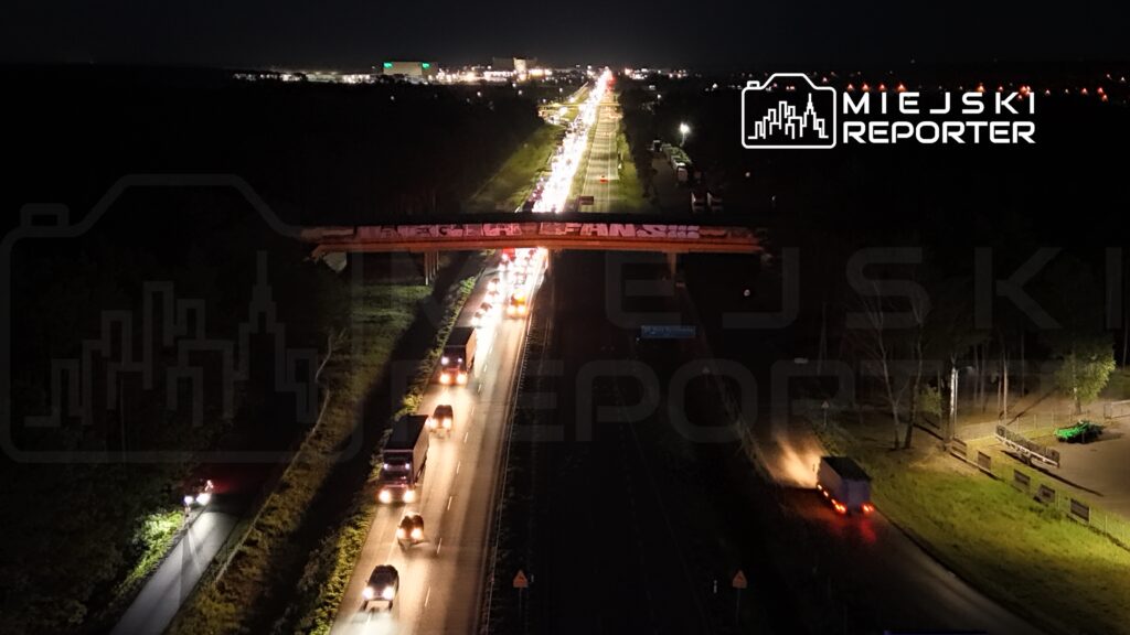 Ruch samochodowy na drodze w nocy, z widocznym mostem z graffiti w tle oraz oświetleniem ulicznym.