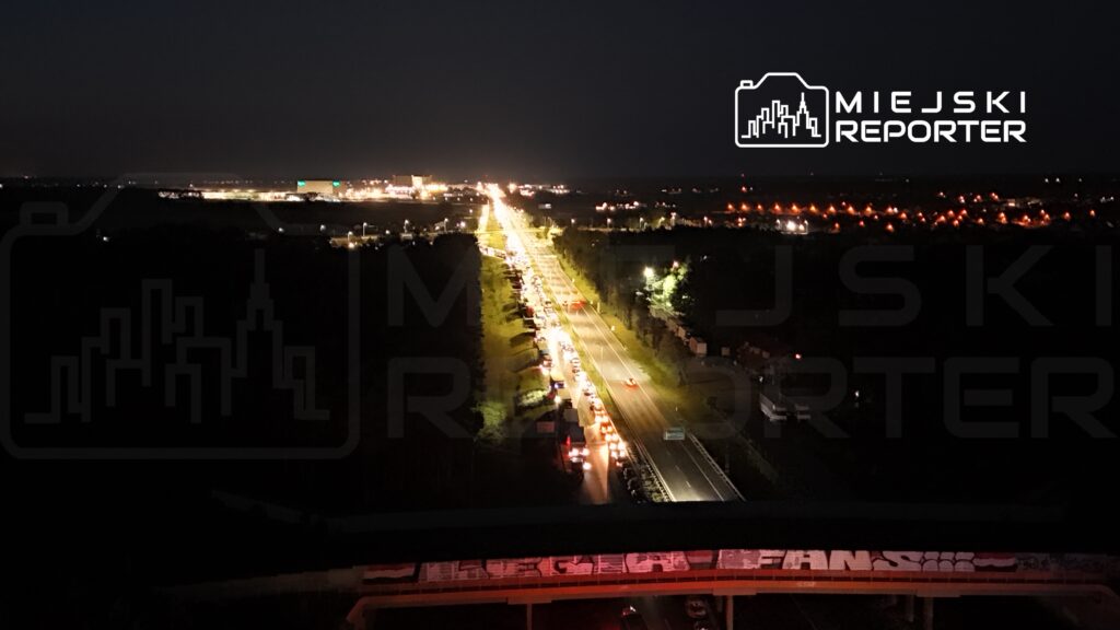 Ruch samochodowy na oświetlonej drodze w nocy, z długim sznurkiem pojazdów w kolejce. W tle widać iluminację budynków.