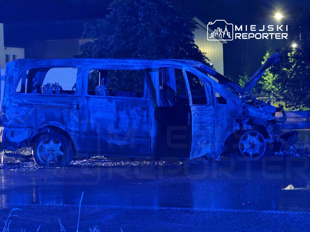 Spalony pojazd dostawczy z otwartymi drzwiami, stojący na mokrej nawierzchni nocą, oświetlony niebieskimi światłami.