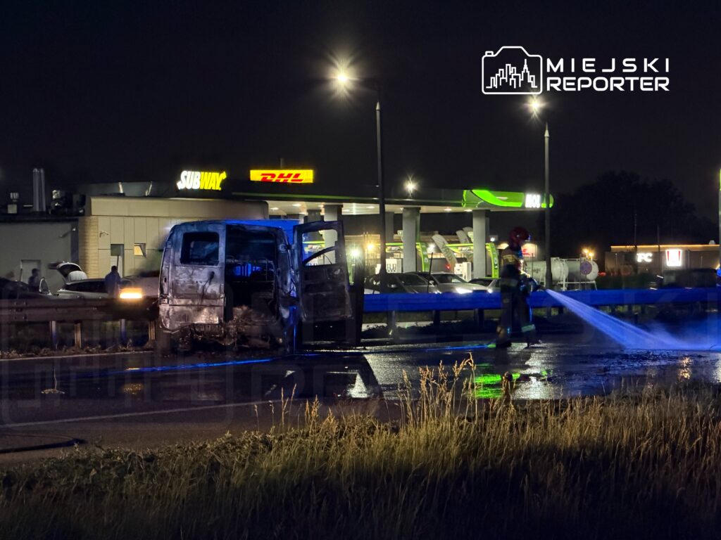 Strażak gasi płonącego furgona przy drodze, obok stacji benzynowej i fast fooda, w nocnej scenerii.