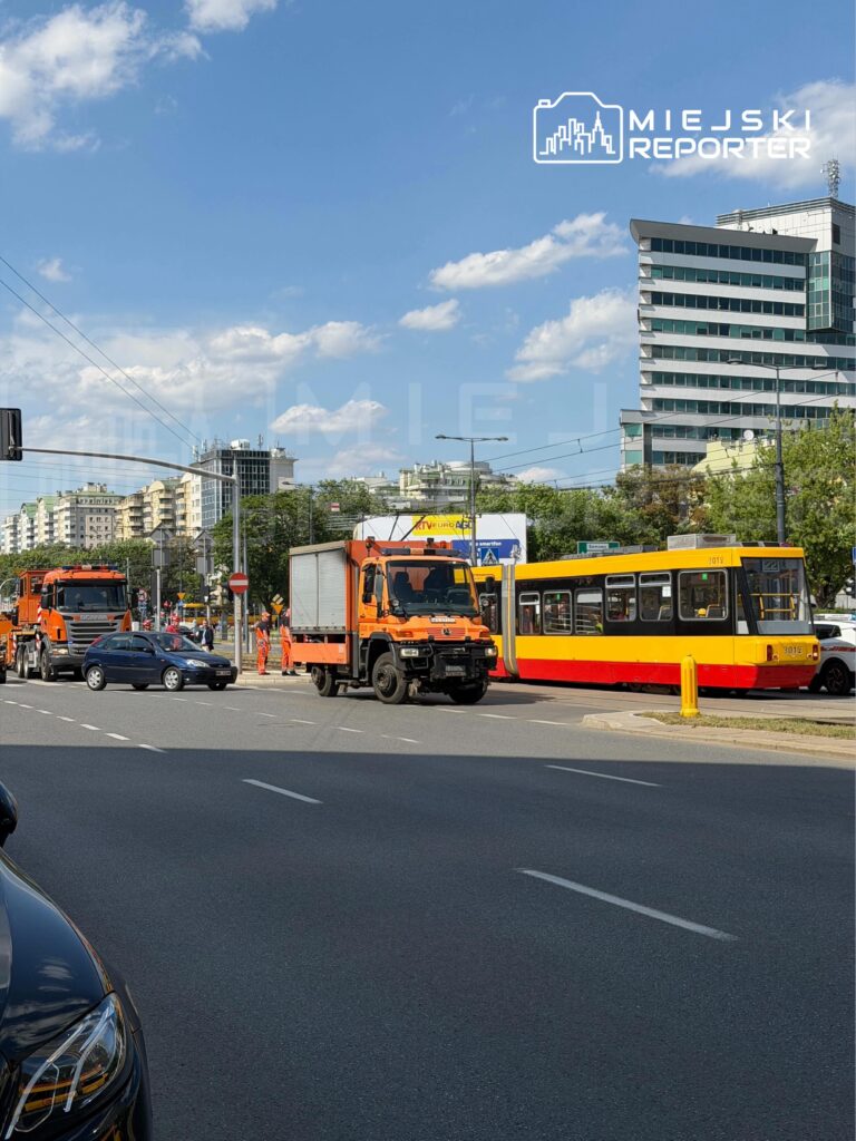 Pomarańczowy pojazd dostawczy stoi na skrzyżowaniu, obok tramwaju z czerwono-żółtym malowaniem, w miejskim otoczeniu.