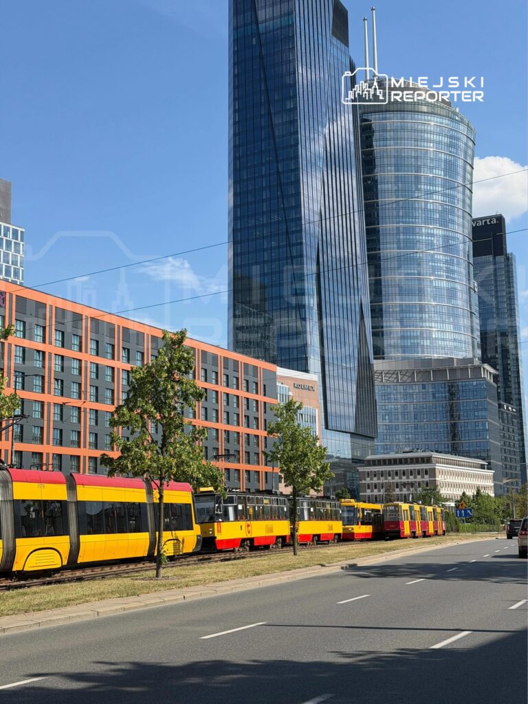 Tramwaje w żółto-czerwonych kolorach stoją na torach obok nowoczesnych biurowców w miejskim otoczeniu.