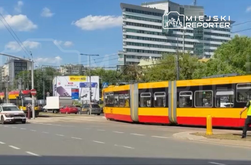 Tramwaj w kolorze żółto-czerwonym skręca na skrzyżowaniu, obok ruchliwego pasa drogowego z samochodami. W tle nowoczesny biurowiec.
