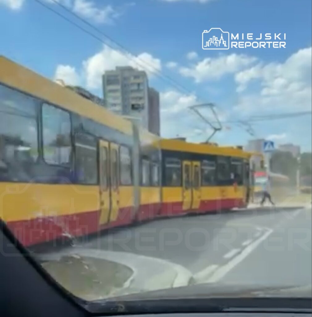 Tramwaj w żółto-czerwonych barwach skręca w prawo na zakręcie, w tle widoczny nowoczesny budynek i niebieskie niebo.