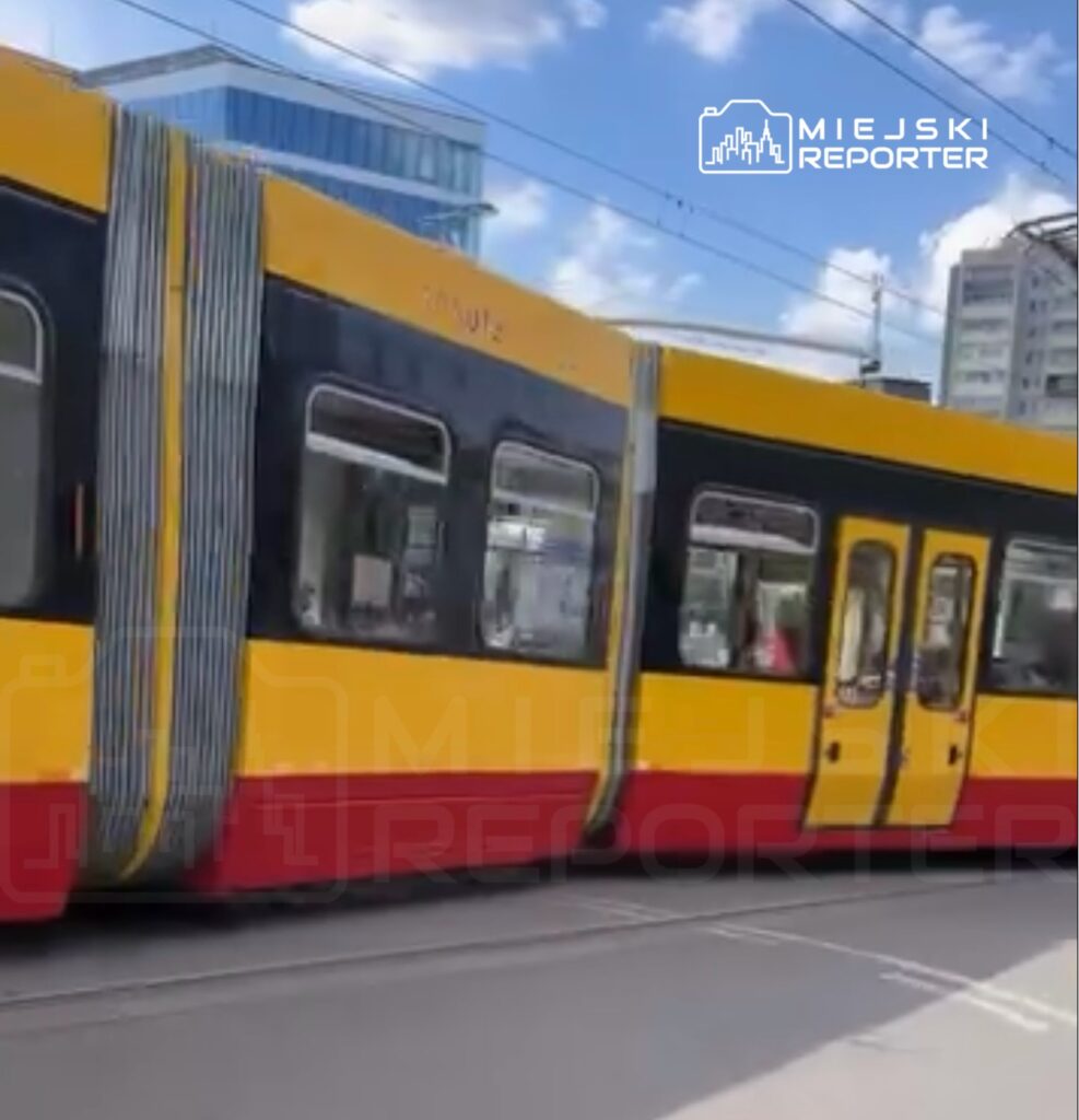 Tramwaj w żółto-czarnych barwach przejeżdża po torach w miejskim otoczeniu, w tle nowoczesne budynki.