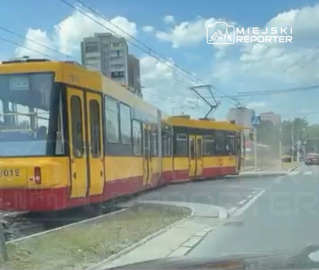 Tramwaj w żółto-czerwonych barwach skręca na skrzyżowaniu w miejskiej okolicy, obok wysokich budynków.