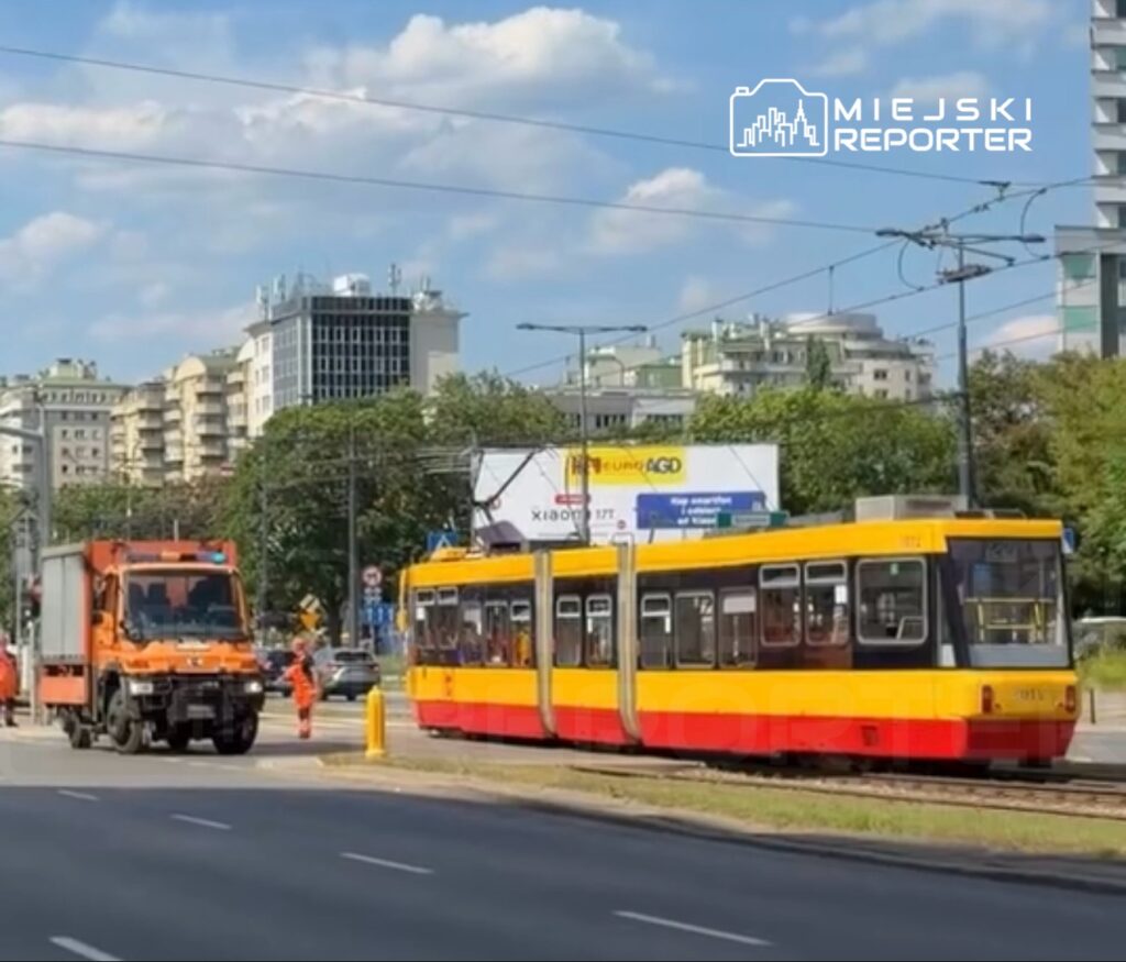 Tramwaj w żółto-czerwonym malowaniu zatrzymany na przystanku, obok pomarańczowy pojazd służb komunalnych.