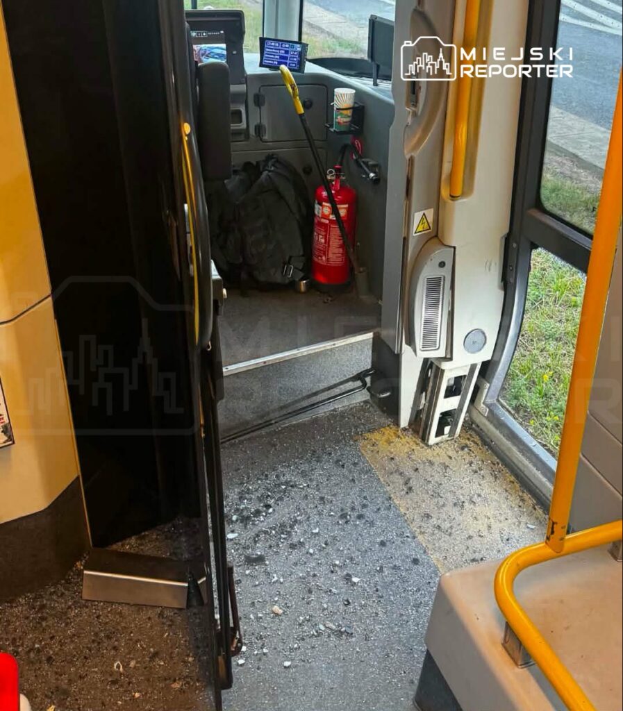 Wnętrze autobusu z widoczną rozlaną substancją na podłodze, obok drzwi znajduje się gaśnica i plecak.