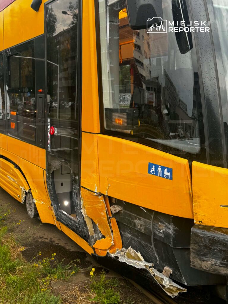 Uszkodzony tramwaj z wgniecioną boczną częścią na przystanku, w deszczowej aurze, obok ruchliwej ulicy.