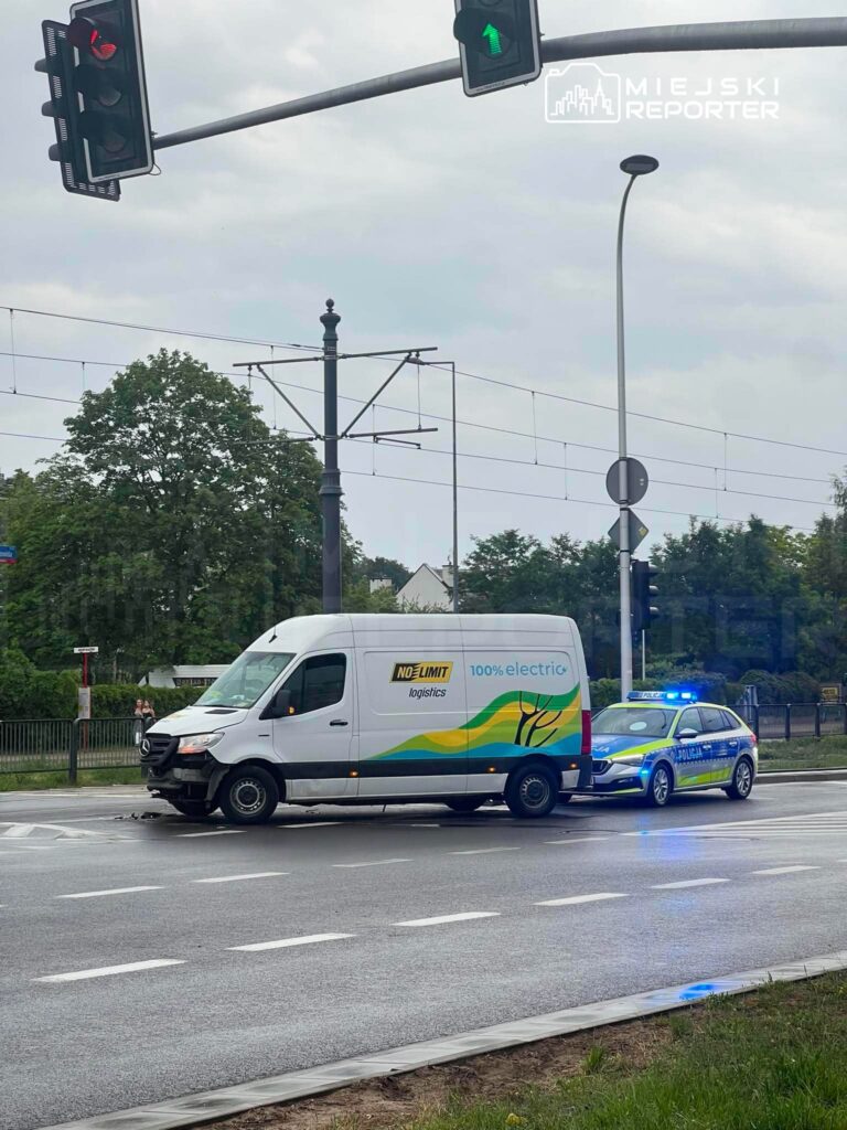 Elektryczna furgonetka zatrzymana przez policyjny radiowóz na skrzyżowaniu, w tle sygnalizacja świetlna.