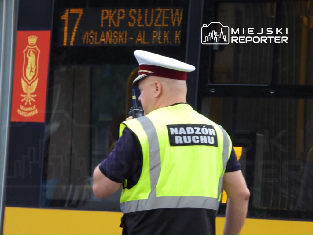 Mężczyzna w kamizelce odblaskowej z napisem "NADZÓR RUCHU" rozmawia przez radio obok tramwaju z wyświetlaczem kierunku.