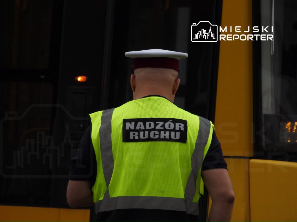 Mężczyzna w kamizelce odblaskowej z napisem "NADZÓR RUCHU" obserwuje ruch przy przystanku komunikacji miejskiej.