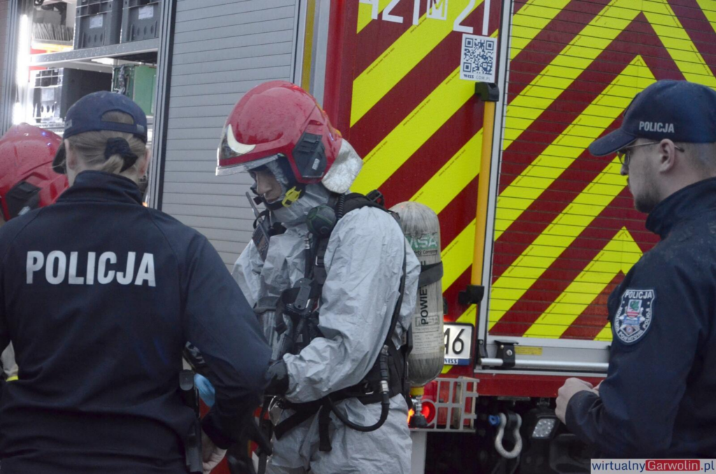 Strażak w kombinezonie ochronnym przygotowuje się do akcji ratunkowej, obok policjantów przy wozie strażackim.