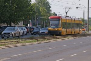 Tramwaj w żółto-czarnych barwach porusza się po torach obok zatoru samochodowego na ulicy otoczonej zielenią.