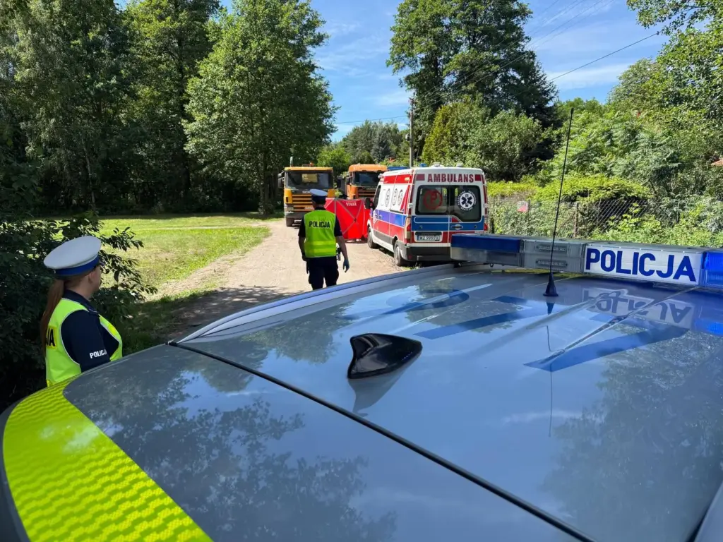 Policjant w kamizelce odblaskowej kieruje ruchem obok policyjnego samochodu, w tle karetka i pojazdy służbowe.