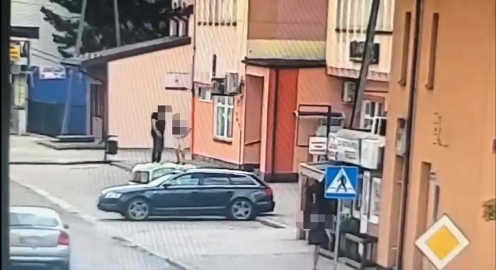 Dwoje ludzi rozmawia na chodniku obok zaparkowanego samochodu w miejskim otoczeniu.