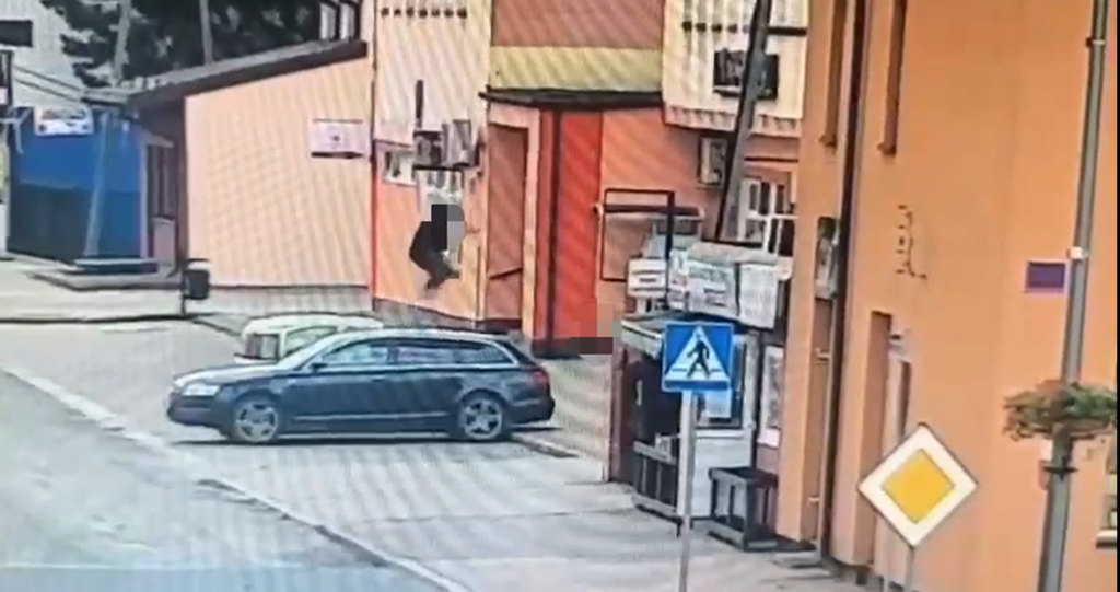 Mężczyzna w ciemnej odzieży schodzi z budynku na ulicy, obok parkujący samochód i znak drogowy.