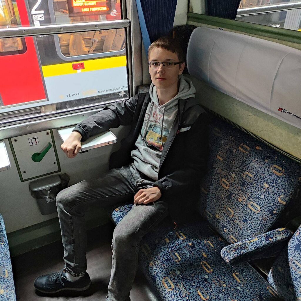 Młody mężczyzna w okularach siedzi na siedzeniu w tramwaju, obok niego widoczny jest okno z widokiem na czerwony tramwaj.