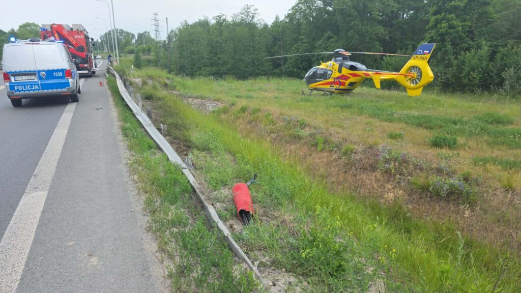 Policyjny radiowóz z niebieskimi światłami stoi na poboczu drogi, obok lądowisko dla helikoptera ratunkowego.
