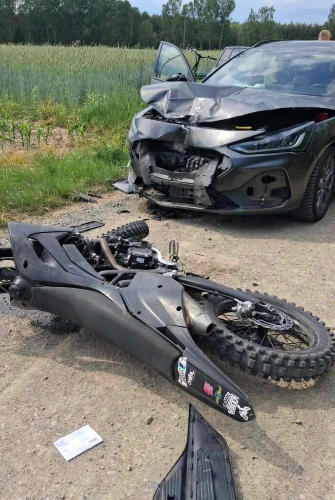 Motocykl leży na drodze obok uszkodzonego samochodu, w tle pola z roślinnością.