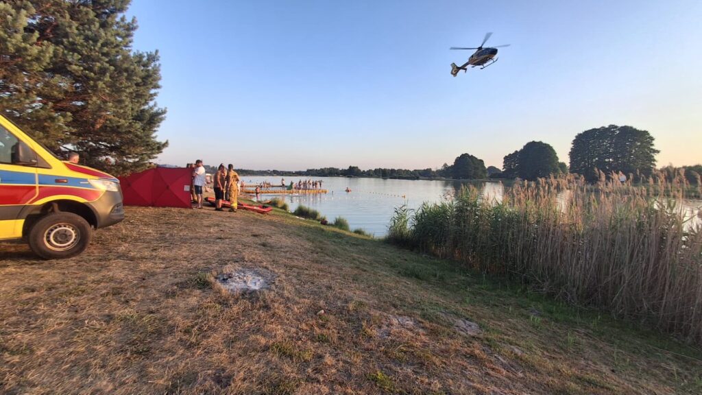 Grupa osób przy brzegu jeziora, obok karetki, podczas akcji ratunkowej z helikopterem w tle.