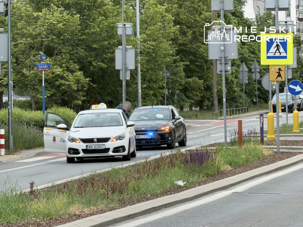 Taxi zatrzymane na drodze, obok policyjny radiowóz z włączonymi sygnałami świetlnymi, w tle zieleń i znaki drogowe.