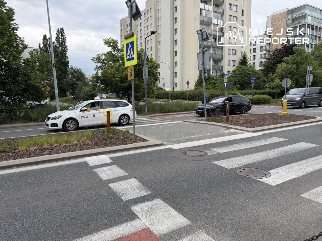 Biały samochód taxi przejeżdża obok przejścia dla pieszych w miejskim otoczeniu z zielenią.