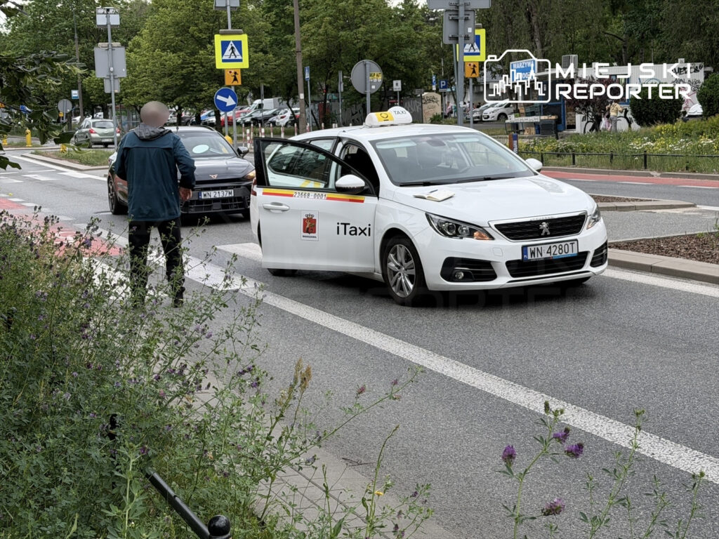 Mężczyzna w zielonej kurtce czeka na przystanku, obok zatrzymane białe taxi na ulicy w miejskim otoczeniu.
